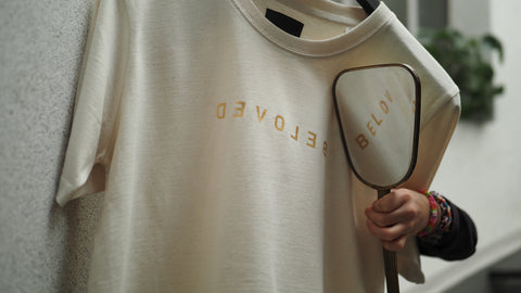 Ein nachhaltiger Beloved Pullover am Kleiderhaken. Ein Handspiegel, spiegelt den Schriftzug BELOVED.